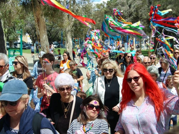Image of A group of people, including children and adults, gather outdoors waving colourful ribbon sticks. They appear to be celebrating or attending a festive event in a park with trees and playground equipment.