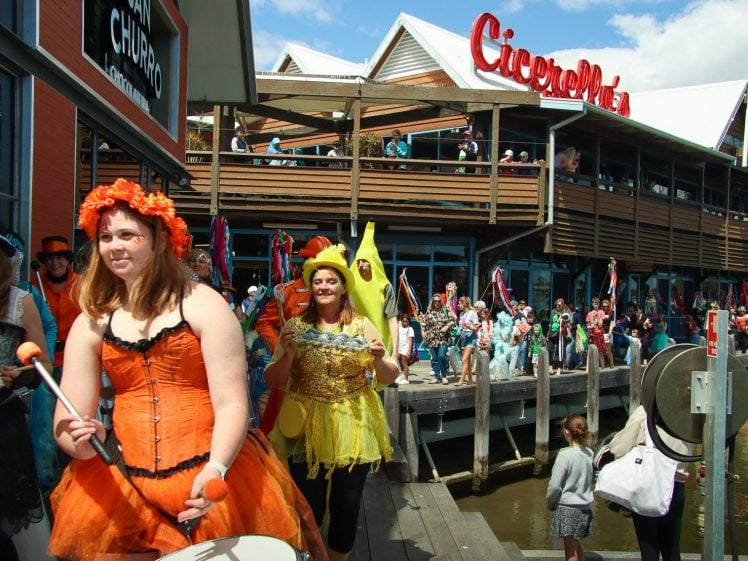 Image of People in colourful costumes, including a woman in an orange dress and another in a yellow outfit, parade and play instruments along a wooden promenade. A crowd watches near a large building labelled Cicerello’s.
