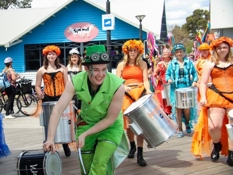 Image of A lively outdoor parade features a person in a bright green costume leading a group of colourfully dressed performers playing drums. The group marches on a wooden boardwalk near a blue building under a sunny sky.