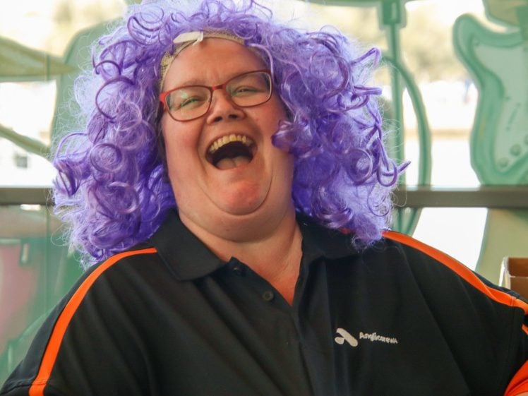 Image of A person wearing glasses and a bright purple curly wig laughs joyfully. They are dressed in a black shirt with orange stripes and are indoors with guitars visible in the background.