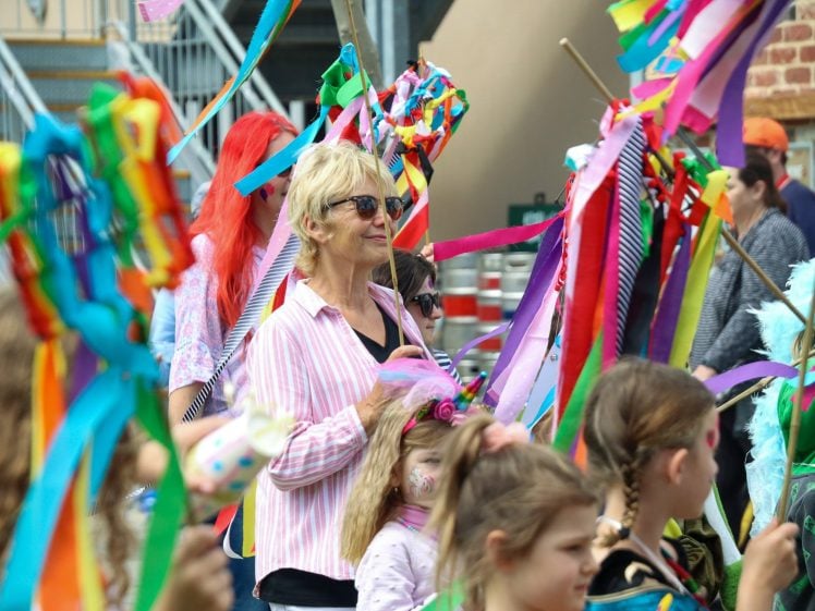 Image of A group of people, including children and adults, participate in an outdoor event holding colourful ribbons and decorated sticks. The atmosphere is festive, with vibrant costumes and face paint visible among the crowd.