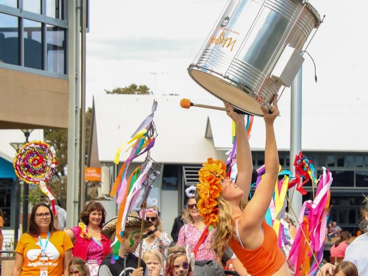 Image of A woman in an orange outfit and flower crown holds a large drum above her head whilst performing at a colourful outdoor parade, surrounded by people with costumes, face paint, and ribbon wands.