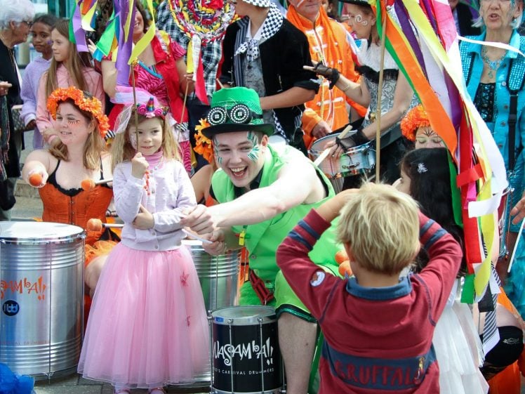 Image of A lively group of people in colourful costumes, including adults and children, celebrate outdoors with music and laughter. Some wear fancy hats, bright dresses, and face paint. A boy faces the camera amid the festive crowd.