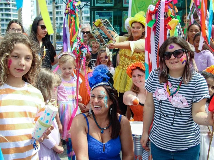 Image of A lively group of children and adults in colourful costumes and face paint celebrate outdoors with ribbons, musical instruments, and bright decorations. The atmosphere is festive and joyful.