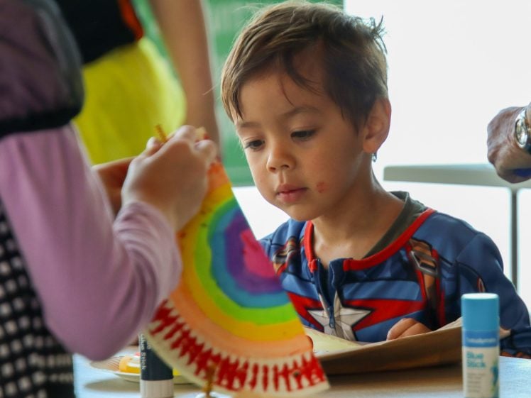 Image of A young child in a superhero costume looks intently at a colourful rainbow craft held by another person, with glue sticks and art supplies visible on the table.