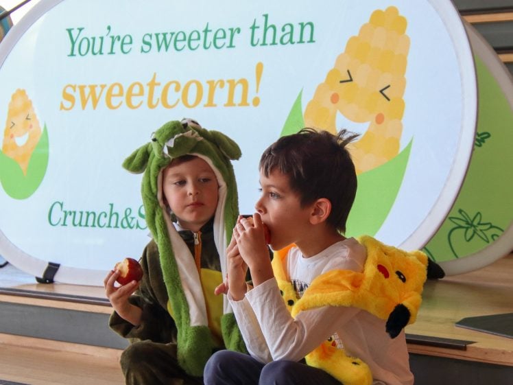 Image of Two children in animal costumes sit on steps eating apples. Behind them is a sign with a smiling corn image and text that reads, You’re sweeter than sweetcorn! Crunch&.