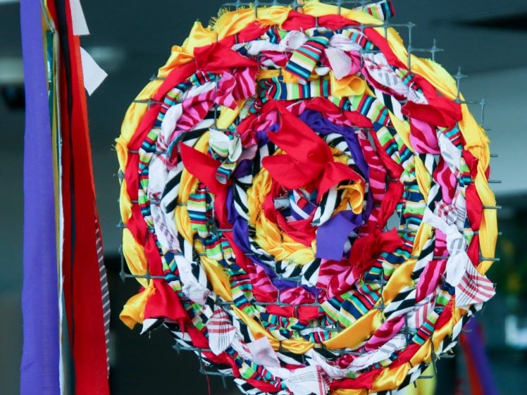 Image of A large, colourful circular decoration made of twisted and braided strips of fabric in red, yellow, purple, and patterned colours, resembling a lollipop or swirl, with long ribbons hanging from the side.