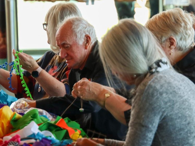 Image of Four older adults sit at a table, working with colourful fabric and decorations. They are focused on crafting, using bright materials in a well-lit indoor space.