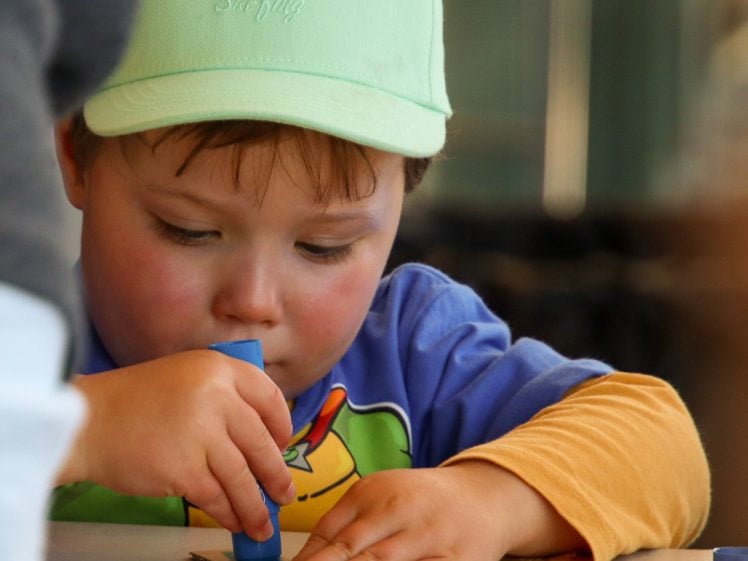 Image of A young child wearing a light green cap and colourful shirt concentrates whilst colouring a piece of cardboard with a blue crayon at a table.