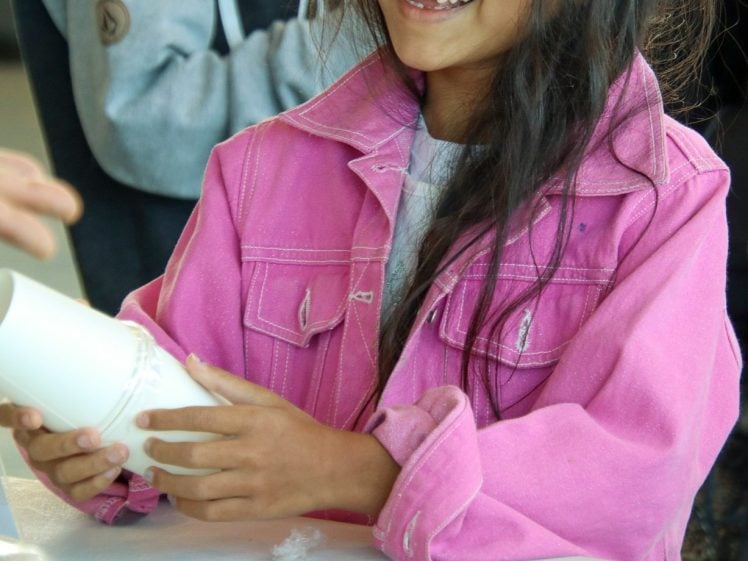 Image of A smiling young girl in a pink jacket sits at a table holding a white paper tube, surrounded by craft supplies like tape, scissors, pencils, and a paper plate. Another child is visible in the background.