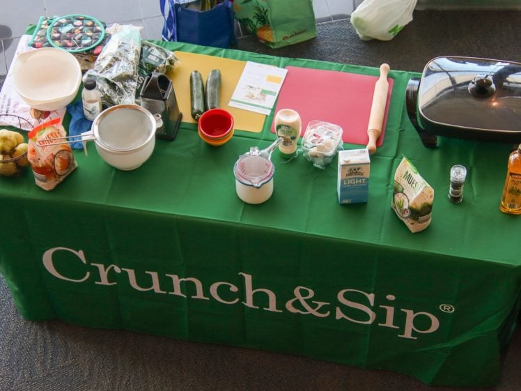 Image of Overhead view of a green table with Crunch&Sip written on the cloth, set up with various cooking ingredients, utensils, and an electric grill, ready for a cookery demonstration.