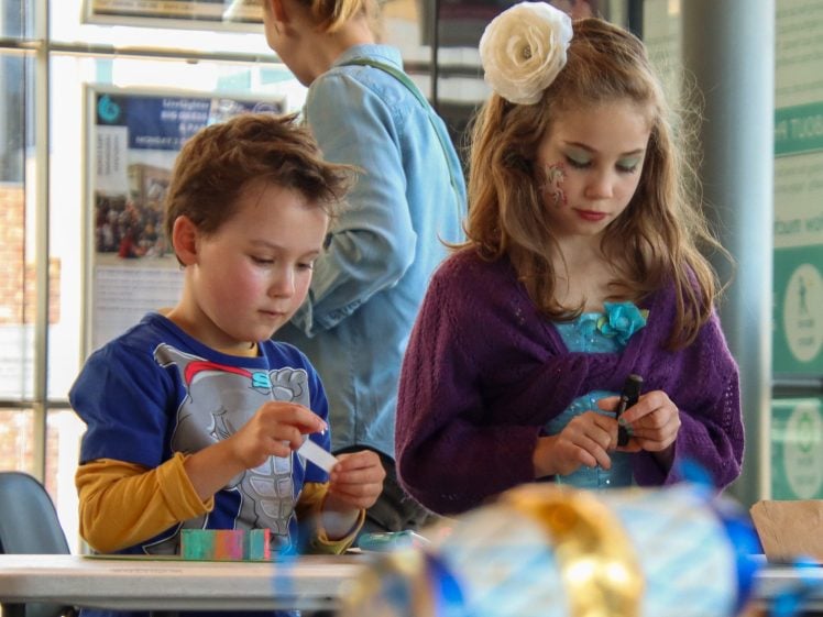 Image of Two young children in fancy dress work on crafts at a table. The boy wears a superhero outfit, and the girl has a flower headband and face paint. An adult stands in the background, slightly out of focus.
