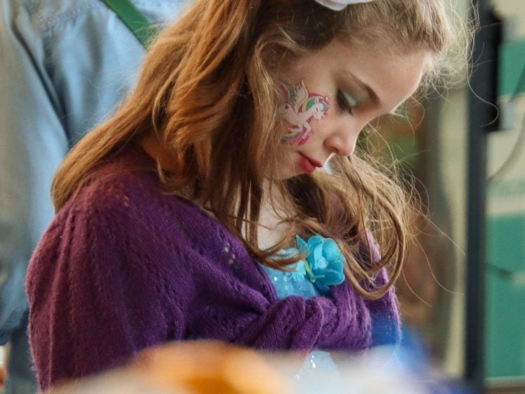 Image of A young girl with a white flower hair accessory and face paint looks down thoughtfully. She wears a purple jumper and has wavy brown hair. A blurred person and objects are in the background and foreground.