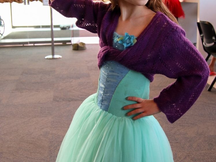 Image of A young girl in a mint green dress and purple jumper poses indoors with one hand on her hip and the other raised, holding a small object. She has a white flower in her hair and face paint on her cheek.