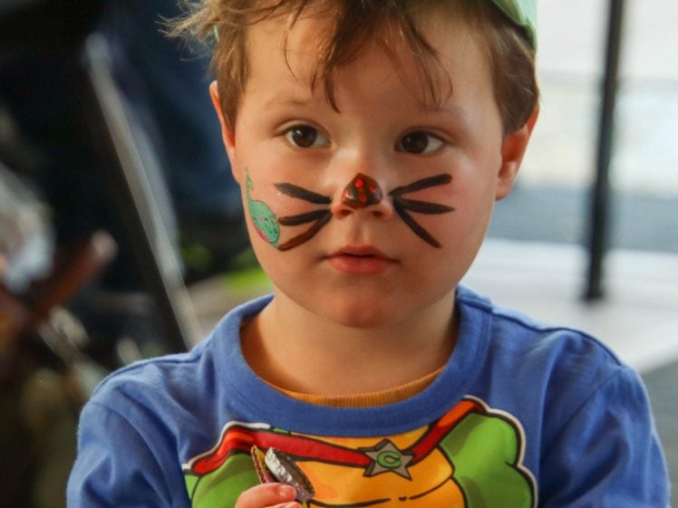 Image of A young child with face paint resembling a cat, wearing a green cap and a blue shirt with a cartoon character, holds a snack whilst looking slightly to the side.
