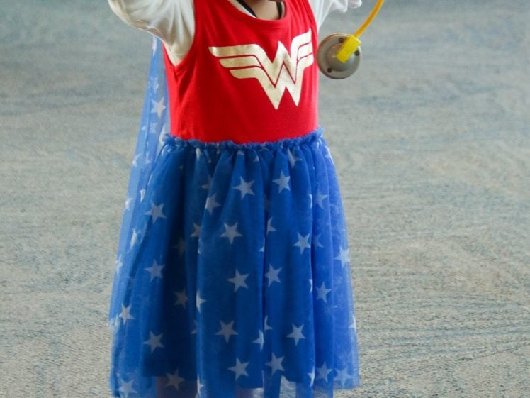 Image of A young child in a Wonder Woman costume and white trainers holds a toy stethoscope to her head while standing indoors, with people blurred in the background.
