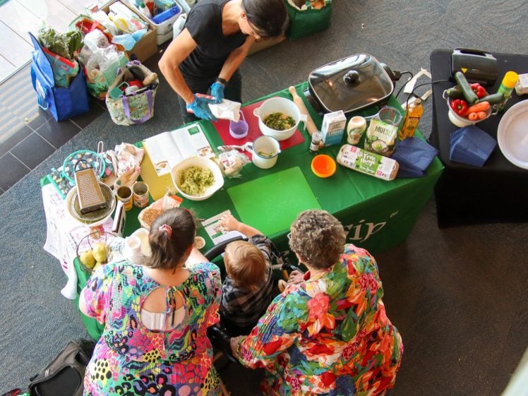 Image of Overhead view of a cookery demonstration, with a chef preparing food at a green table while two women in colourful dresses and a child watch. Shopping bags and food items are scattered around the area.