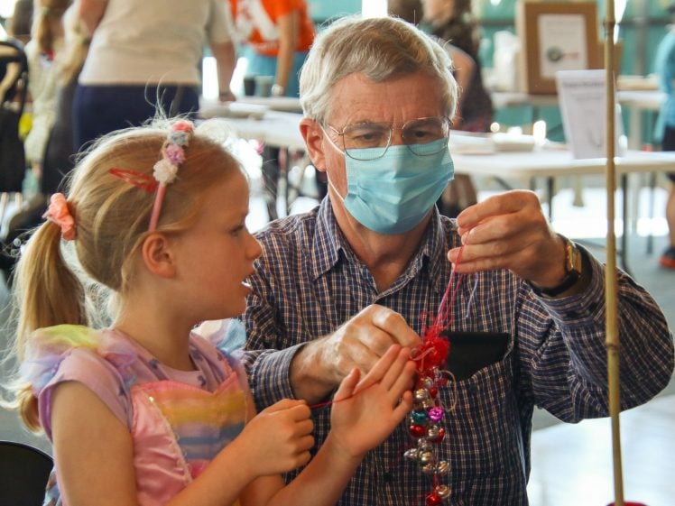 Image of An older man wearing a mask helps a young girl in a rainbow dress with a craft project, threading red wool together at a table in a busy indoor setting.