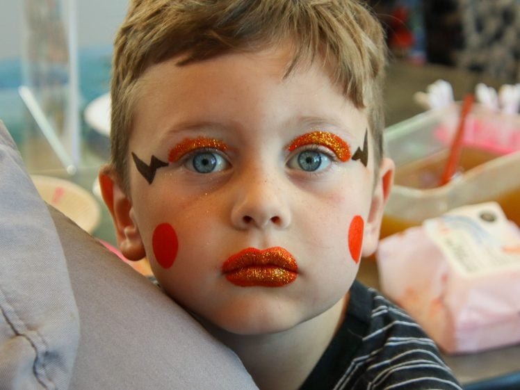 Image of A young boy with short light brown hair and blue eyes has his face painted with red cheeks, glittery red lips, and bold orange and black eye make-up. He is looking directly at the camera, wearing a striped shirt.