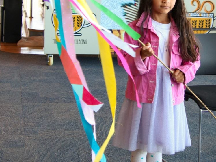 Image of A young girl in a pink jacket and white dress holds a stick decorated with colourful ribbons, standing indoors on a carpeted floor with a chair and creative signs in the background.