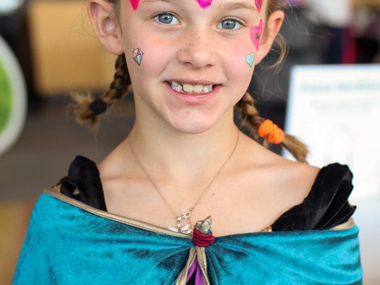 Image of A smiling young girl with plaited hair and bright face paint, including pink hearts and blue details, wearing a turquoise cape over a fancy-dress costume, standing indoors.