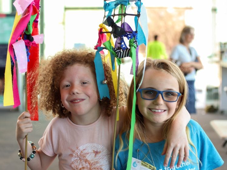 Image of Two children smile and pose together indoors, holding colourful handmade crafts with ribbons and bells. One child has curly hair and the other wears glasses. Bright sunlight streams through windows in the background.