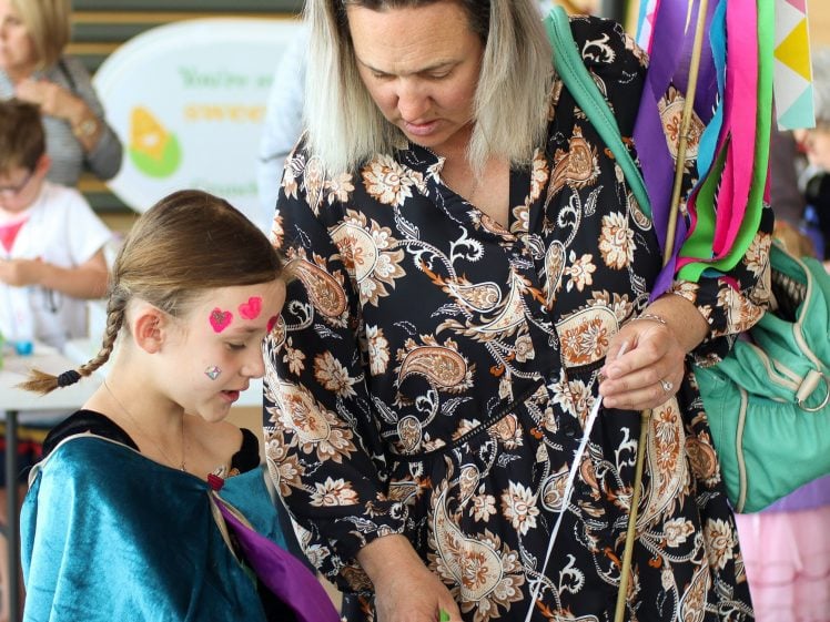 Image of A woman in a floral dress helps a girl with face paint and heart stickers with an art activity. Colourful ribbons and other children are visible in the background at the event.