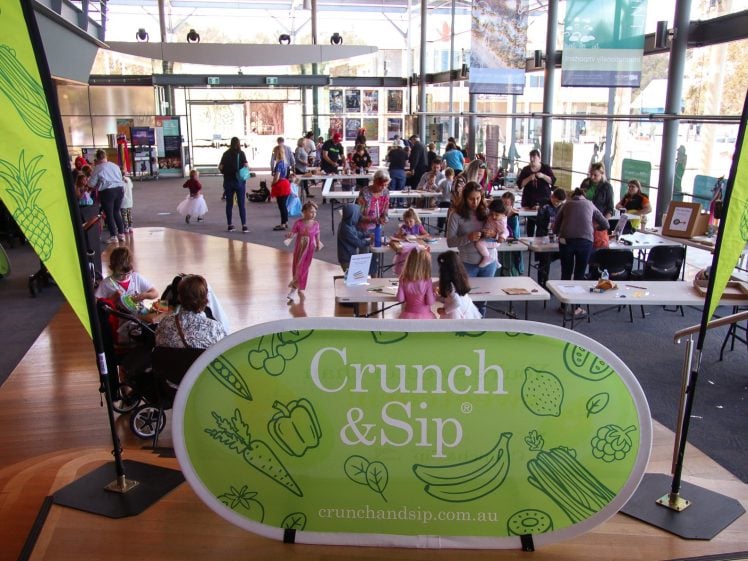 Image of People gather in a bright indoor venue for a community event. A green Crunch & Sip sign and banners are in the foreground, whilst families and children participate in activities at tables in the background.