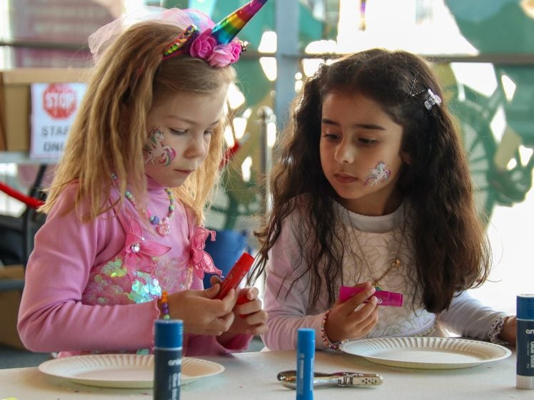 Image of Two young girls with face paint and colourful outfits sit at a table with craft supplies, concentrating on making something. One wears a unicorn headband; both have paper plates and glue sticks in front of them.