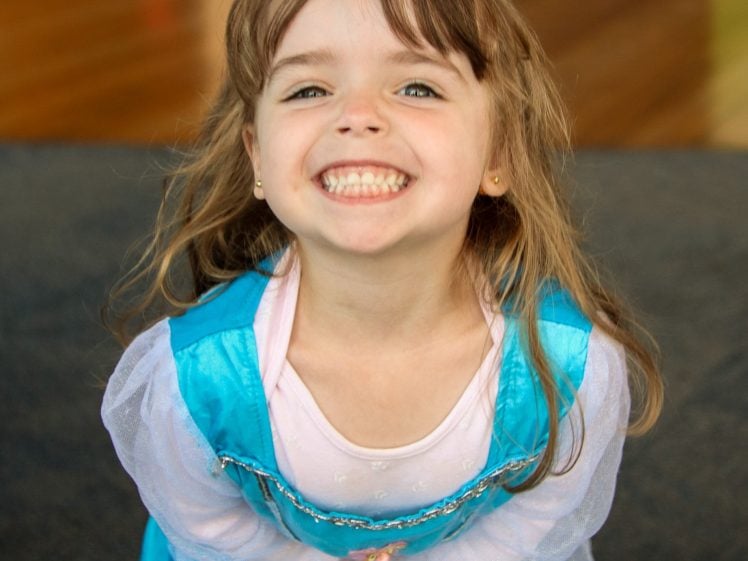 Image of A young girl with long brown hair smiles widely, showing her teeth. She is wearing a light blue princess dress over a pink shirt and standing indoors on a carpeted floor.