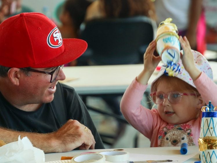 Image of A man in a red San Francisco 49ers cap smiles at a young girl with glasses who is holding a decorated paper craft above her head. They are sitting at a table with more craft supplies and decorations.