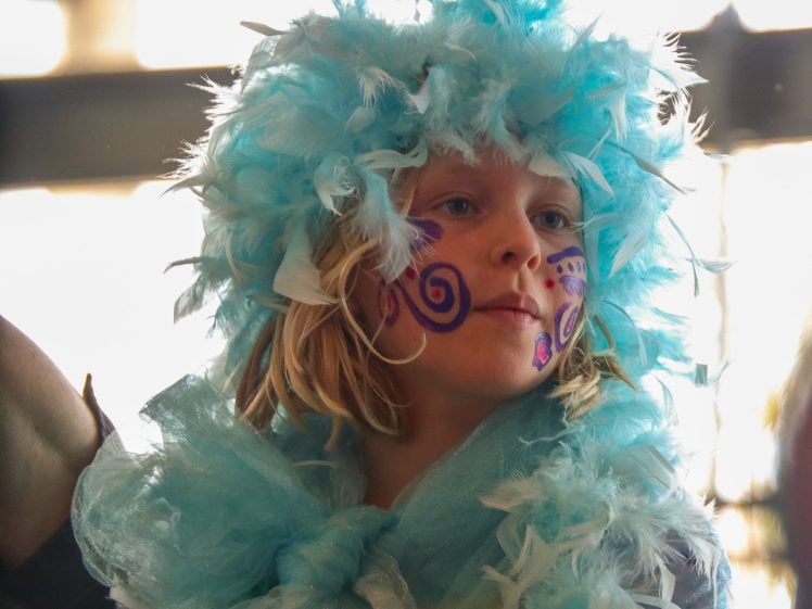 Image of A child with face paint and purple swirls wears a fluffy blue feathered headdress and matching scarf, looking upwards with a thoughtful expression.