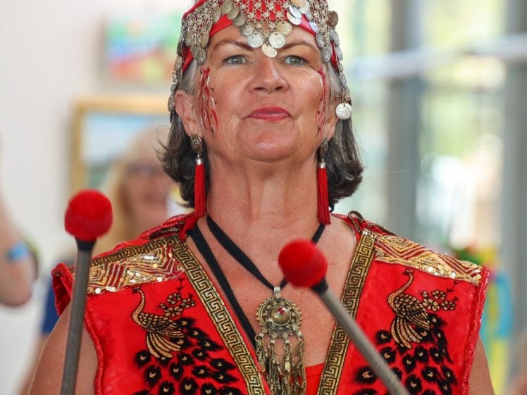 Image of A woman wearing a red, beaded costume with elaborate headpiece and earrings holds two mallets with red tips. She has face paint and stands indoors, with blurred people and art in the background.