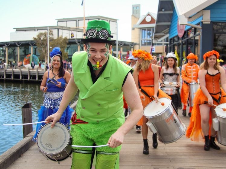 Image of A man in a bright green costume leads a group of drummers dressed in orange and colourful outfits along a promenade near water, smiling and playing music during a festive outdoor parade.