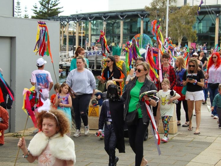 Image of A lively outdoor parade with adults and children dressed in colourful costumes, waving bright, ribboned sticks and marching along a walkway. Spectators watch and join the cheerful procession on a cloudy day.