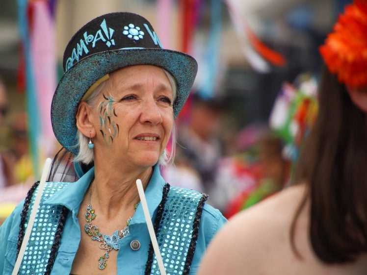Image of A woman in a decorated top hat and blue sequinned outfit participates in a parade, holding drumsticks. She has face paint and wears a blue necklace, with colourful, blurred figures in the background.