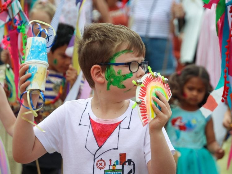 Image of A young boy with green face paint wears glasses and a white shirt with a science design, holding colourful handmade items at a lively outdoor event with other children in fancy dress and decorations.