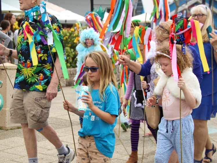Image of A group of people, including children and adults, walk in a parade holding colourful ribbon sticks. Some wear festive clothing and accessories, while others carry drinks. The atmosphere is lively and celebratory outdoors.