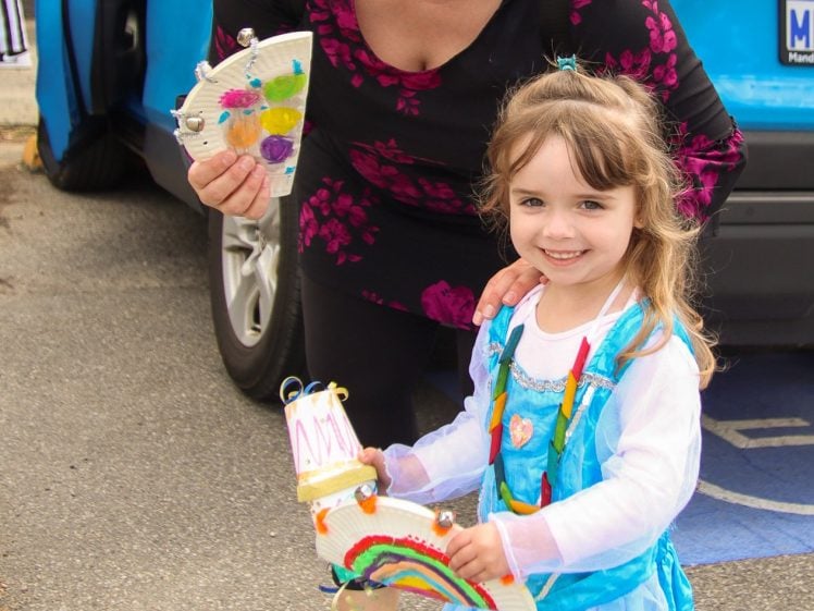 Image of A woman in sunglasses and a black floral dress smiles with a young girl in a blue princess costume. They hold colourful crafts in front of a blue car in a car park. Both look happy and cheerful.