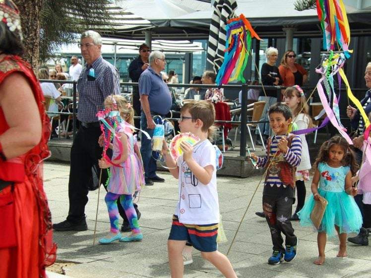 Image of A group of children holding colourful ribbons and decorations walk in a festive outdoor parade, accompanied by adults. People sit at tables in the background, and everyone appears to be celebrating.