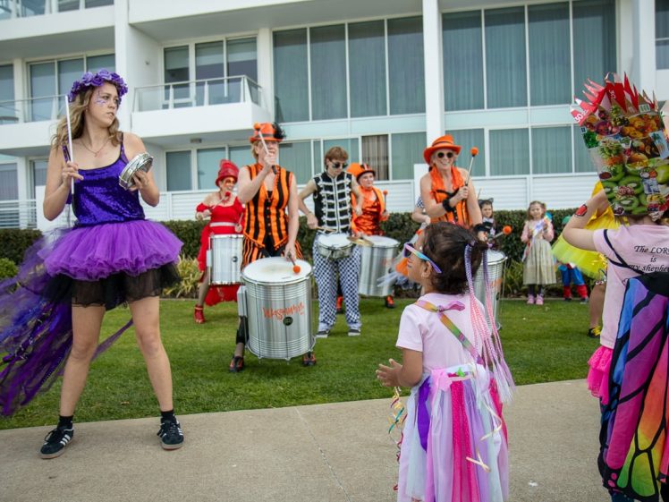 Image of A group of people in colourful costumes perform music outdoors near a modern building, while children in fairy and butterfly costumes watch and interact with them on the grass.