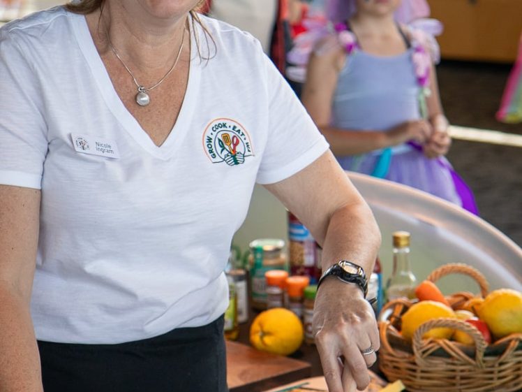 Image of A smiling woman in a colourful sombrero stirs a pan of vegetables at a cookery event. She wears a white shirt with a logo. In the background, a girl in a purple fairy costume stands near a basket of fruit.