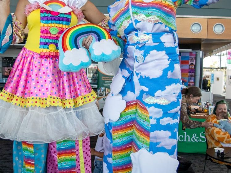 Image of Two people in colourful, whimsical costumes stand on stilts. One wears a rainbow dress with clouds and polka dots, the other wears a sky-themed outfit with clouds and rainbows. Both are smiling and waving indoors.
