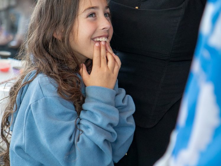 Image of A young girl with long brown hair wearing a blue hoodie smiles with hands near her mouth, looking up excitedly at an adult in a black outfit beside her.