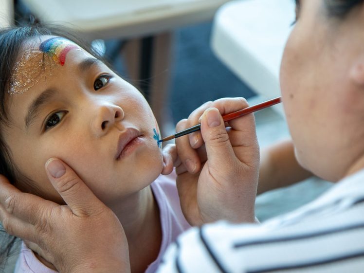 Image of A young child gets her face painted by an adult. She has a small rainbow and glitter on her forehead, whilst the adult carefully adds blue paint to her cheek using a fine brush.