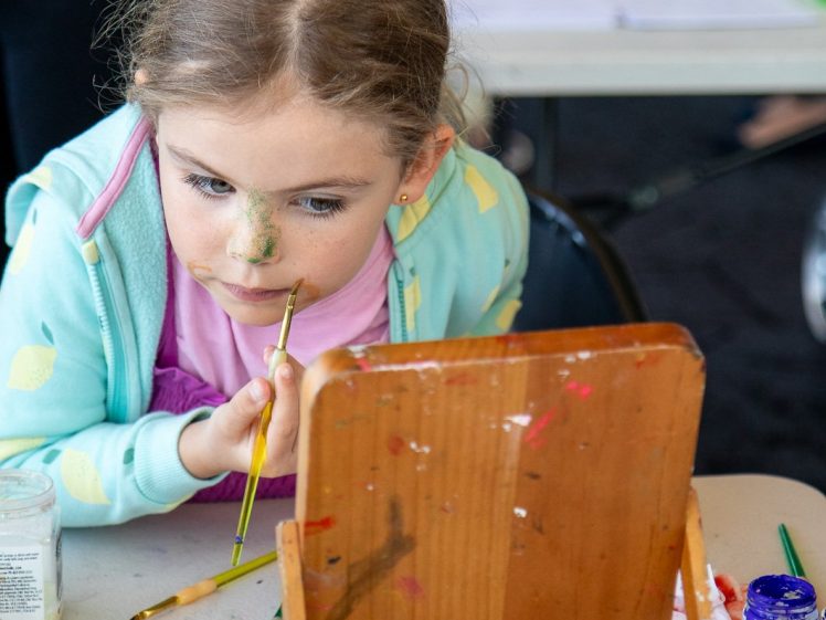 Image of A young girl sits at a table, looking into a wooden mirror whilst painting her nose green with a brush. Art supplies and paint jars are scattered on the table in front of her.
