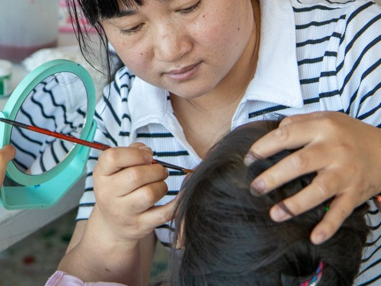 Image of A woman in a striped shirt carefully paints a young girl’s face, gently holding her head steady. The girl has plaited hair with colourful bands and sits patiently, facing away from the camera.