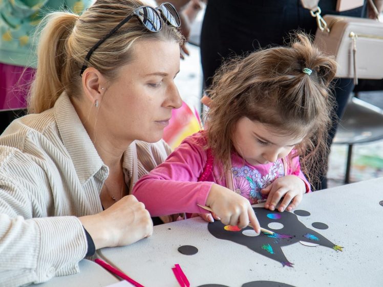 Image of A woman and a young girl sit at a table, focusing on a craft project. The girl decorates a black paper cut-out with colourful dots, while the woman watches and helps. Art supplies are scattered on the table.
