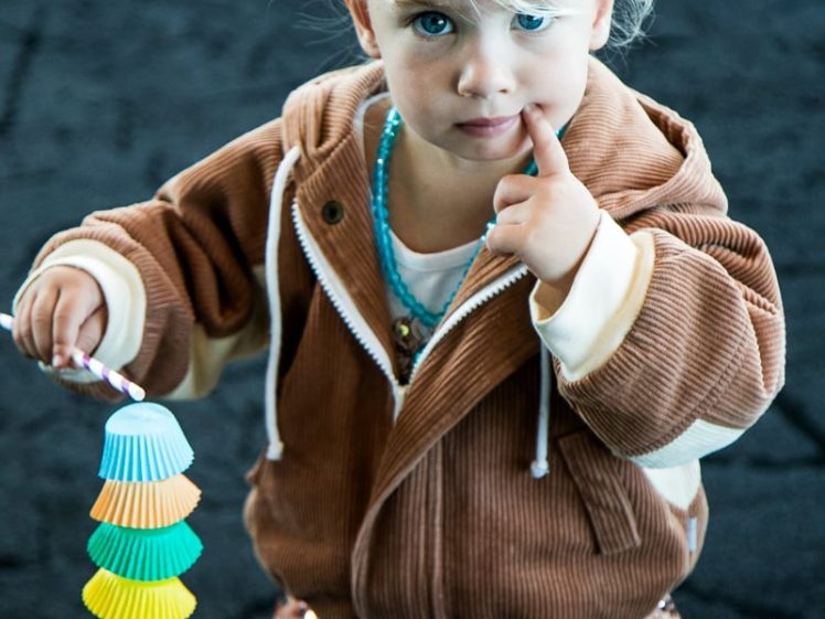 Image of A young child with blonde hair, wearing a brown jacket and a sparkly skirt, holds a colourful toy made of stacked cupcake cases and points with their finger, looking directly at the camera.
