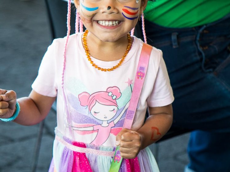 Image of Smiling young girl with face paint, wearing a colourful dress, orange necklace, and carrying a pink handbag. She has a heart painted on her forehead and rainbow lines on her cheeks.